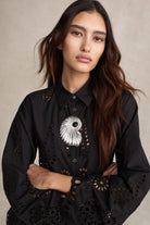 Woman wearing a black blouse with intricate cut-out designs and a decorative brooch.