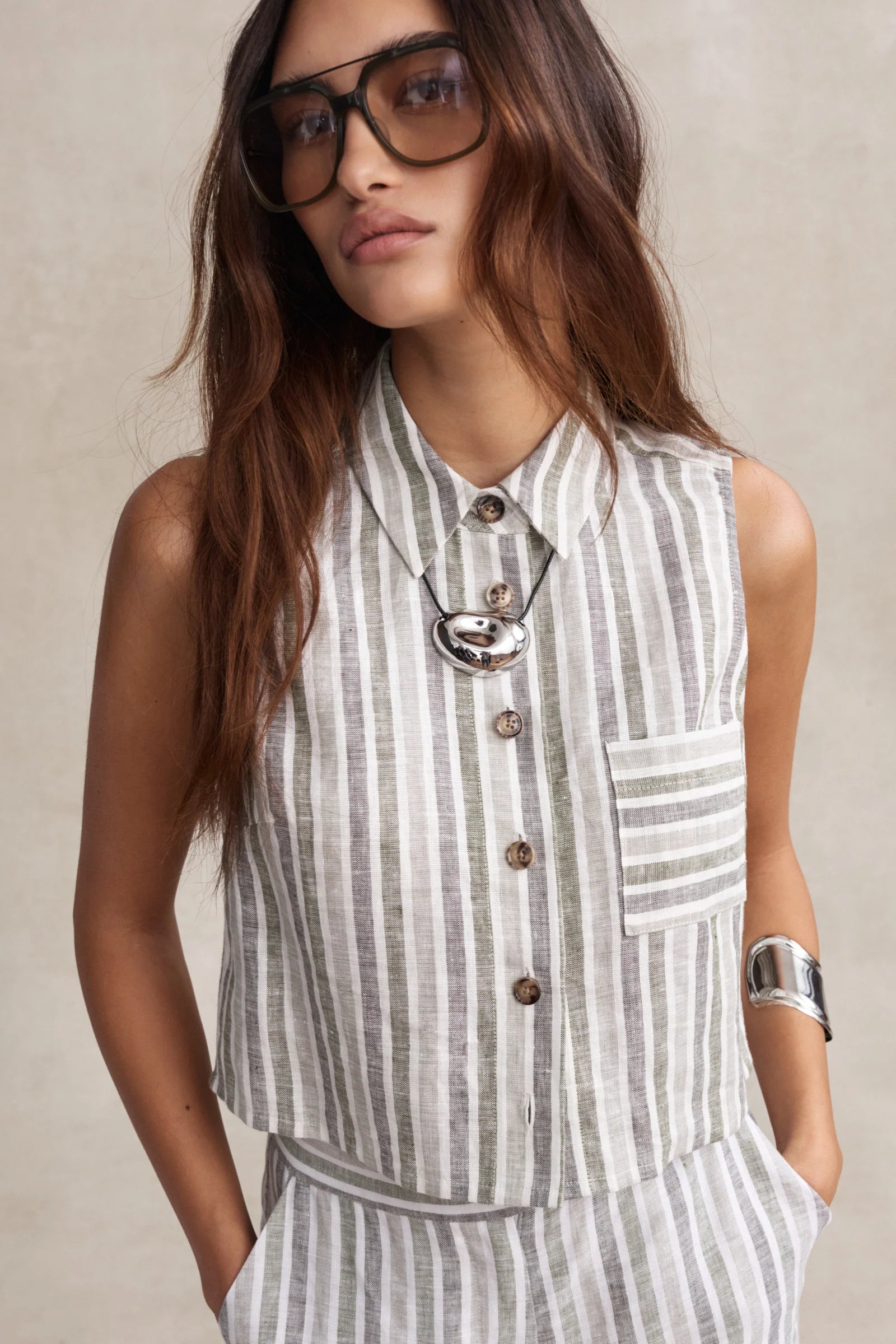 Woman wearing a sleeveless striped top with buttons and a necklace against a neutral background