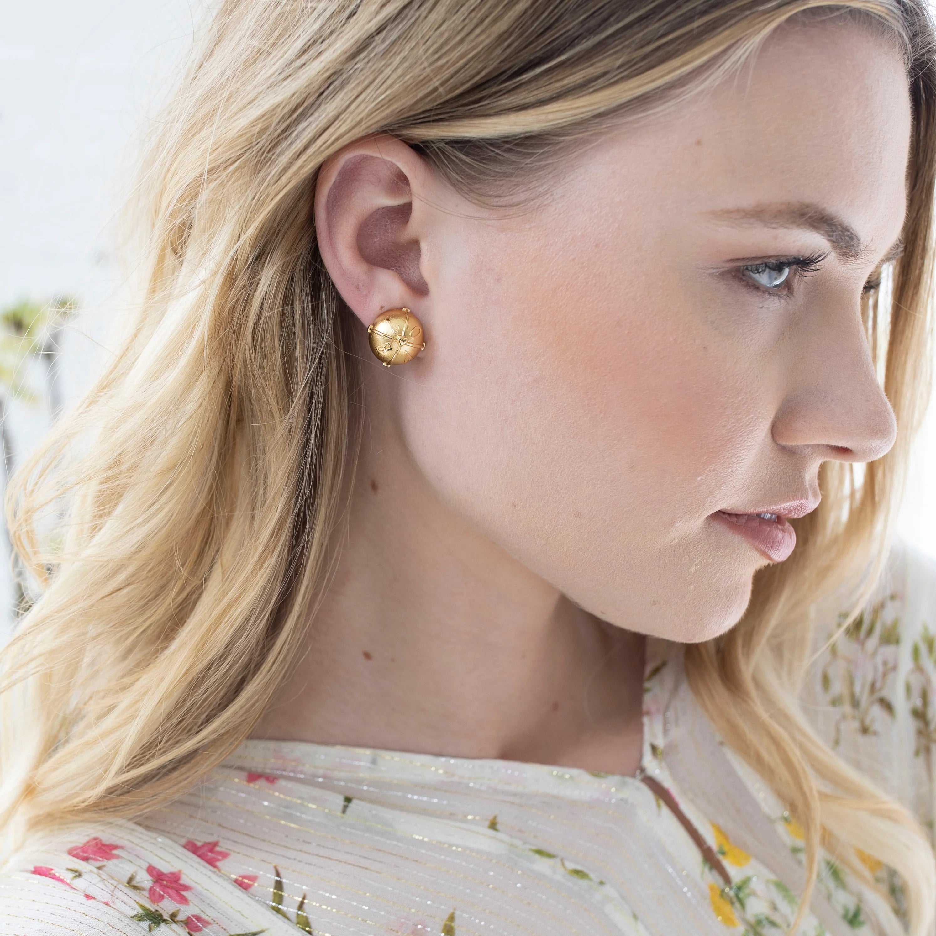 A close-up side profile of a blonde woman wearing a small round gold stud earring with a textured, sunburst-like pattern.