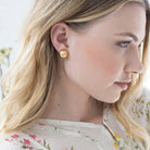 A close-up side profile of a blonde woman wearing a small round gold stud earring with a textured, sunburst-like pattern.