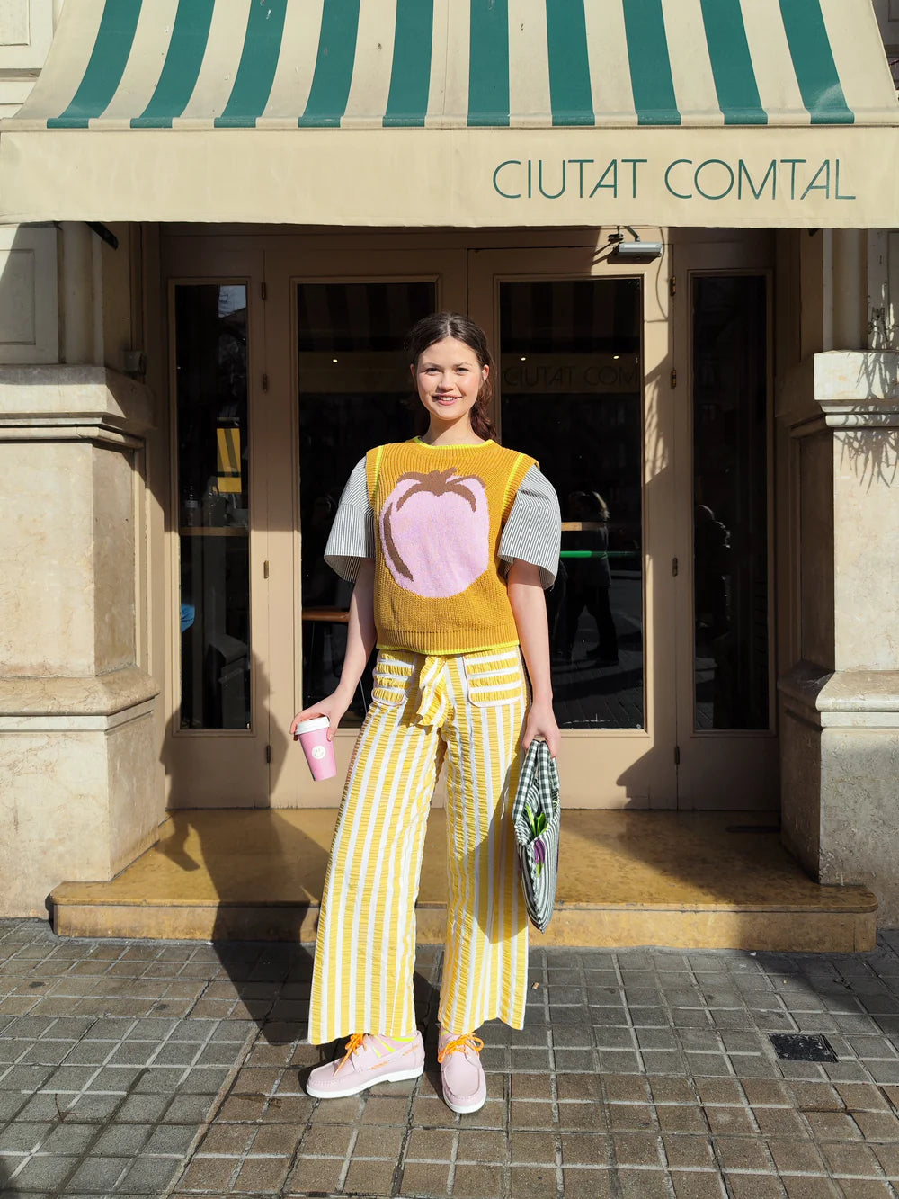 Woman in a mustard yellow top and striped pants standing in front of a building with a green and white striped awning.