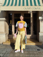 Woman in a mustard yellow top and striped pants standing in front of a building with a green and white striped awning.