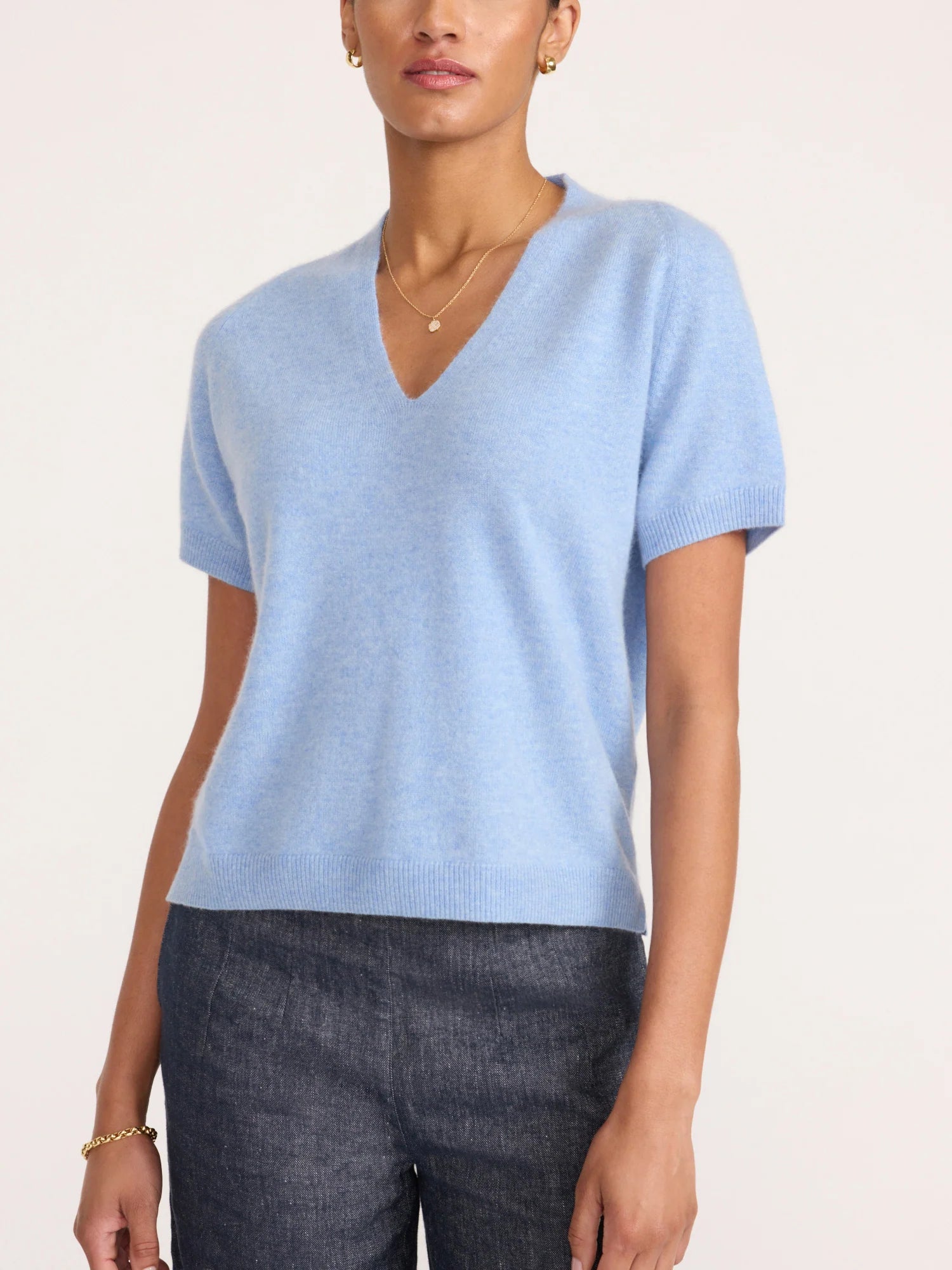A woman wearing a light blue short-sleeve V-neck knit sweater and dark gray jeans, photographed against a plain light background.