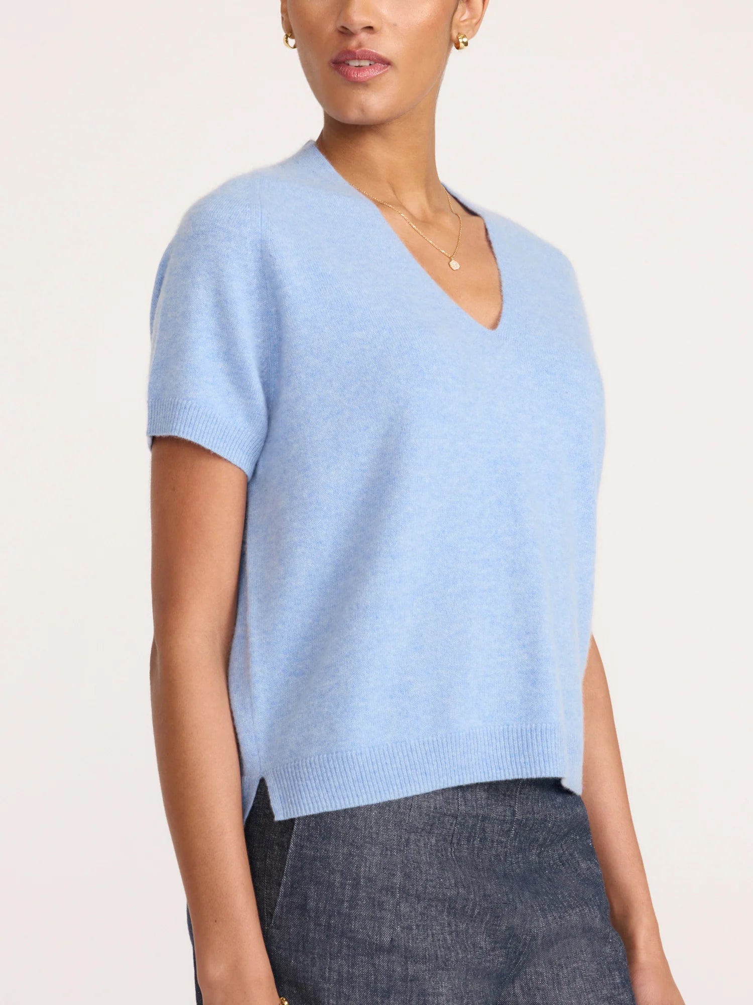 A woman wearing a light blue short-sleeve V-neck knit sweater, paired with dark gray textured trousers, against a plain off-white background.