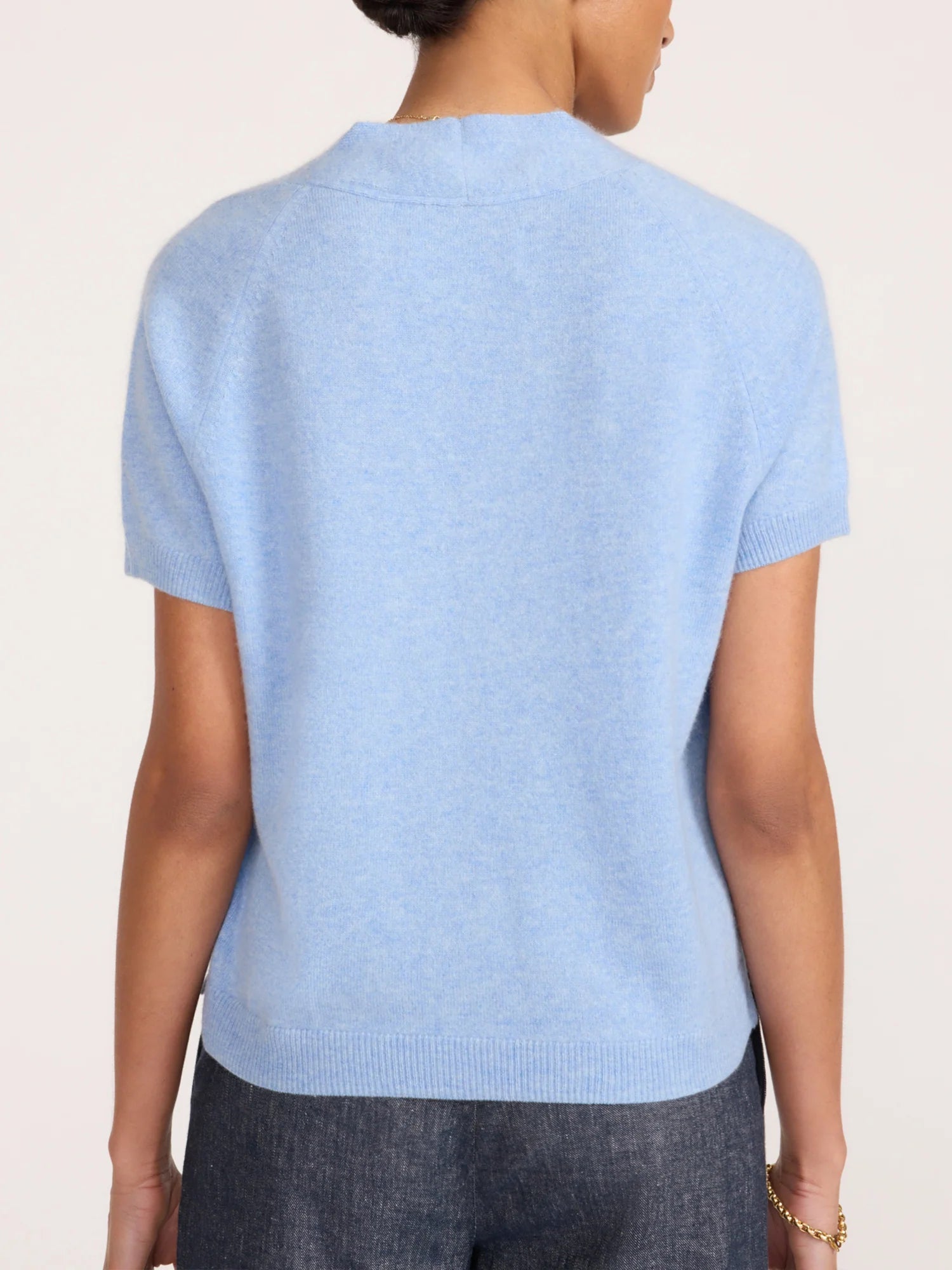 A person shown from the back wearing a light blue short-sleeve knit top and dark gray trousers against a plain light background.