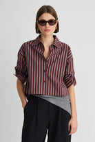 A model wearing a cropped button-up shirt with vertical burgundy and white stripes, styled with a grey undershirt, black trousers, and dark sunglasses against a light gray background.