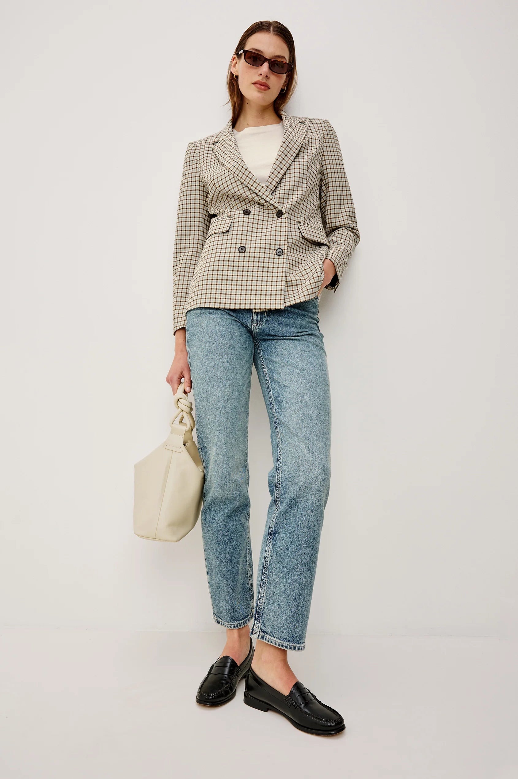Woman wearing a checkered blazer, white shirt, blue jeans, and black shoes on a plain background