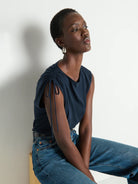 Woman wearing a navy top and blue jeans sitting against a white wall.