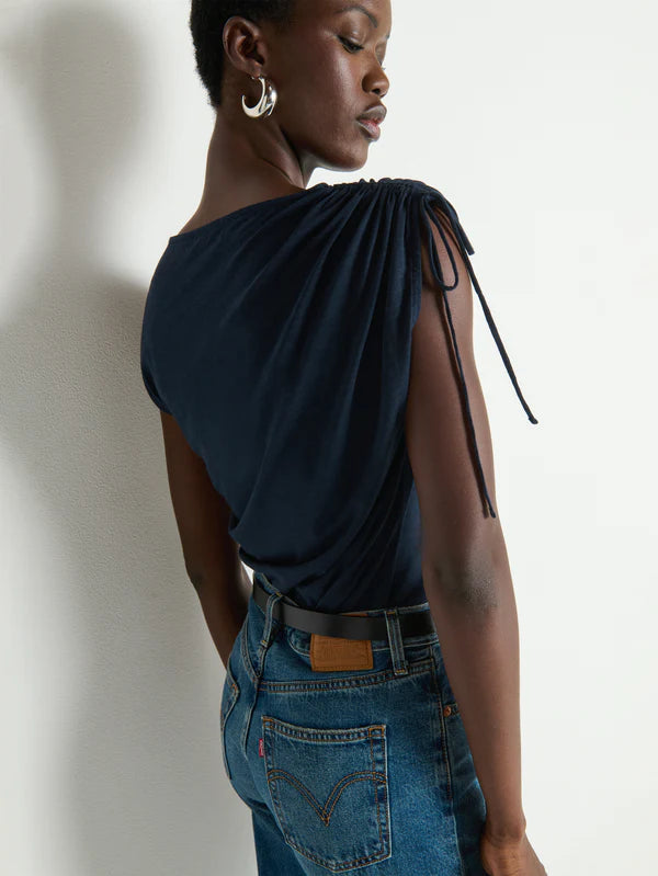 Person wearing a dark blue top with a tie detail and blue jeans against a white wall.
