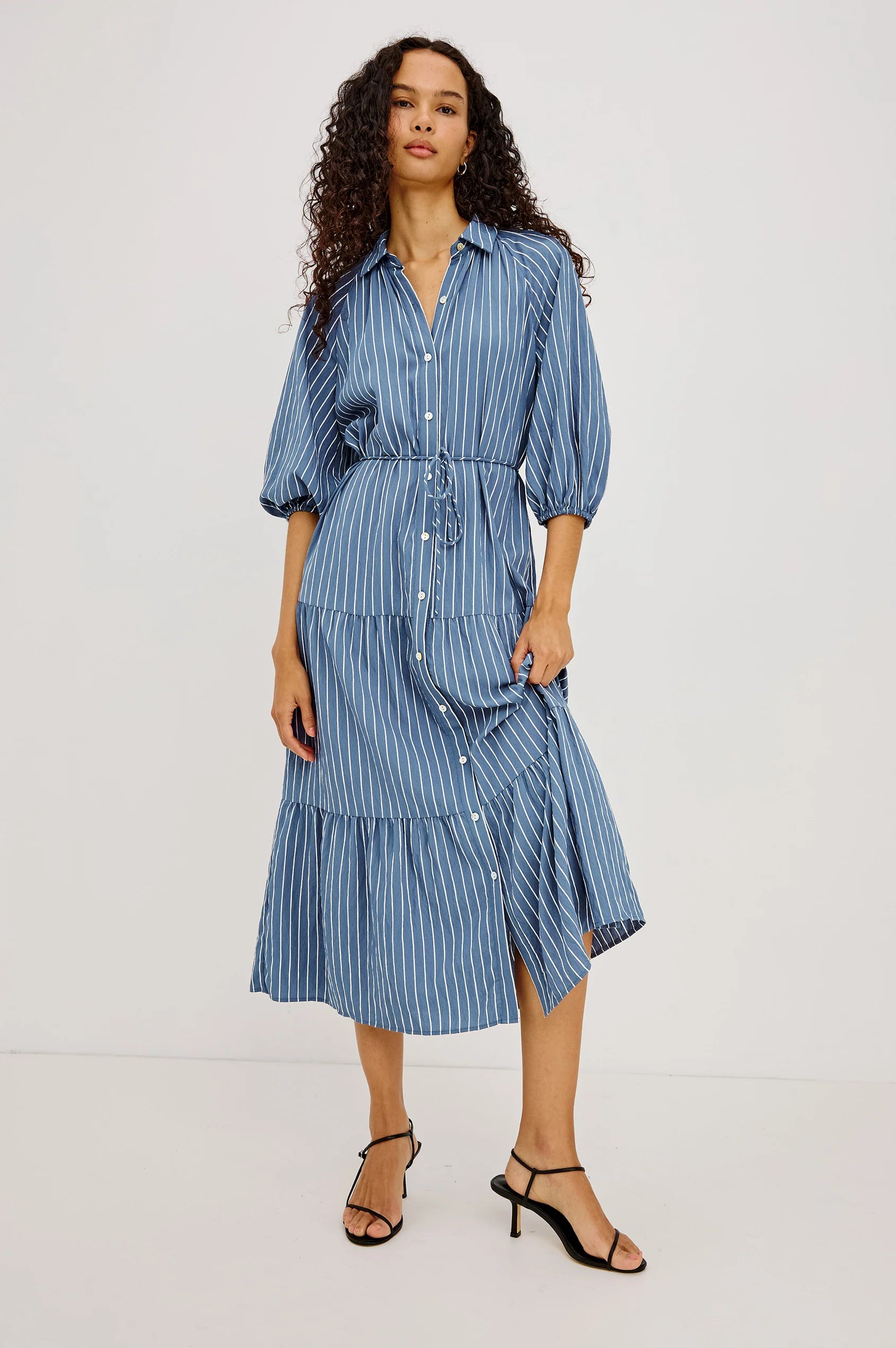 A woman modeling a blue and white vertically striped midi dress with puff sleeves and a tiered skirt, shown from a full body front angle against a plain white studio background.