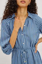 A woman modeling a blue and white vertically striped midi dress with puff sleeves and a tiered skirt, shown from a close up front angle from the mouth to the waist against a plain white studio background.