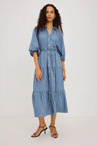 A woman modeling a blue and white vertically striped midi shirt dress with puff sleeves, a button-up front, and a tiered skirt, paired with black strappy heels against a plain light gray studio background.