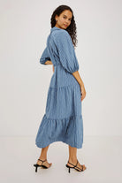 A woman modeling a blue and white vertically striped midi dress with puff sleeves and a tiered skirt, shown from a three-quarter back angle against a plain white studio background.