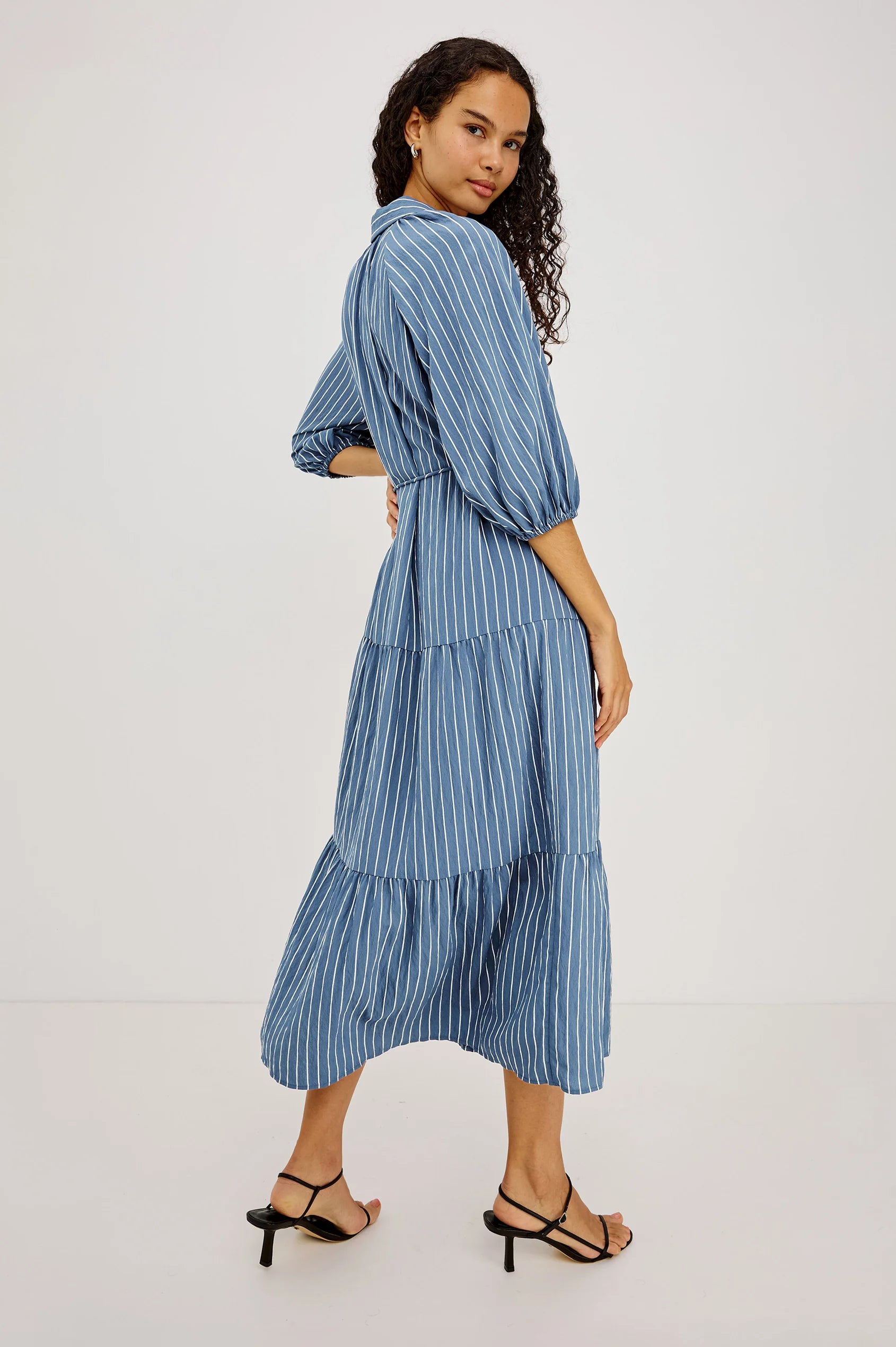 A woman modeling a blue and white vertically striped midi dress with puff sleeves and a tiered skirt, shown from a three-quarter back angle against a plain white studio background.