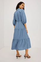 A woman modeling a blue and white vertically striped midi dress with puff sleeves and a tiered skirt, shown from a full body back angle against a plain white studio background.