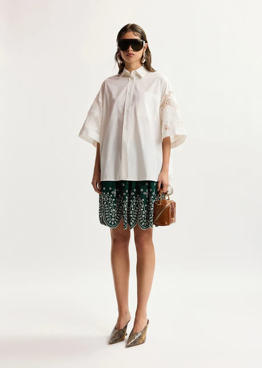 A model stands against a plain light background wearing an oversized white short-sleeve button-up shirt with floral embroidery on the sleeves, dark green and white patterned skirted shorts, pointed metallic heels, and carrying a small brown handbag.
