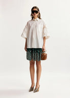 A model stands against a plain light background wearing an oversized white short-sleeve button-up shirt with floral embroidery on the sleeves, dark green and white patterned skirted shorts, pointed metallic heels, and carrying a small brown handbag.