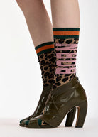 A close-up of a person's legs wearing olive green peep-toe high-heeled ankle boots and patterned socks with leopard print, green and orange stripes, and pink 'MADAM' lettering.