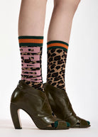 A close-up of a person's legs wearing olive green high-heeled peep-toe shoes and patterned crew socks. One sock features a leopard print pattern with pink lettering, and the other features a pink and black vertical stripe pattern with green and orange stripes at the top.