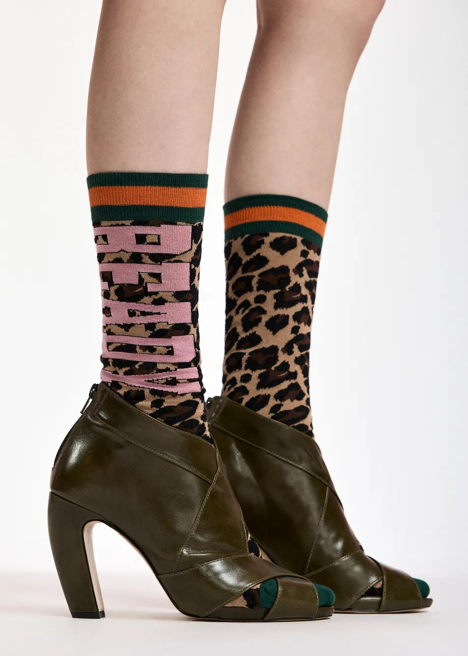 A close-up of a person's legs wearing olive green high-heeled peep-toe shoes and patterned crew socks. One sock features a leopard print pattern with pink lettering, and the other features a pink and black vertical stripe pattern with green and orange stripes at the top.