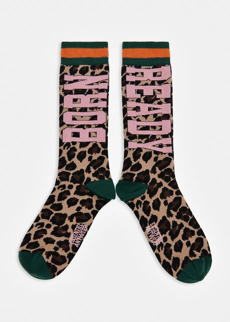 A pair of crew socks with a leopard print pattern, featuring the words 'BORN READY' in pink block letters, dark green heels and toes, and orange and dark green striped cuffs, shown against a plain light gray background.