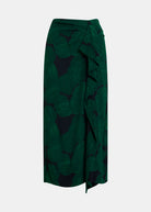 A dark green midi skirt with a tropical leaf print in dark green and black, featuring a ruffled wrap-style waist and side drape, shown against a light gray background.