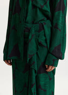 A close-up of a person wearing a long-sleeve belted shirt and matching skirt in a dark green and black abstract pattern, shown against a plain light background.