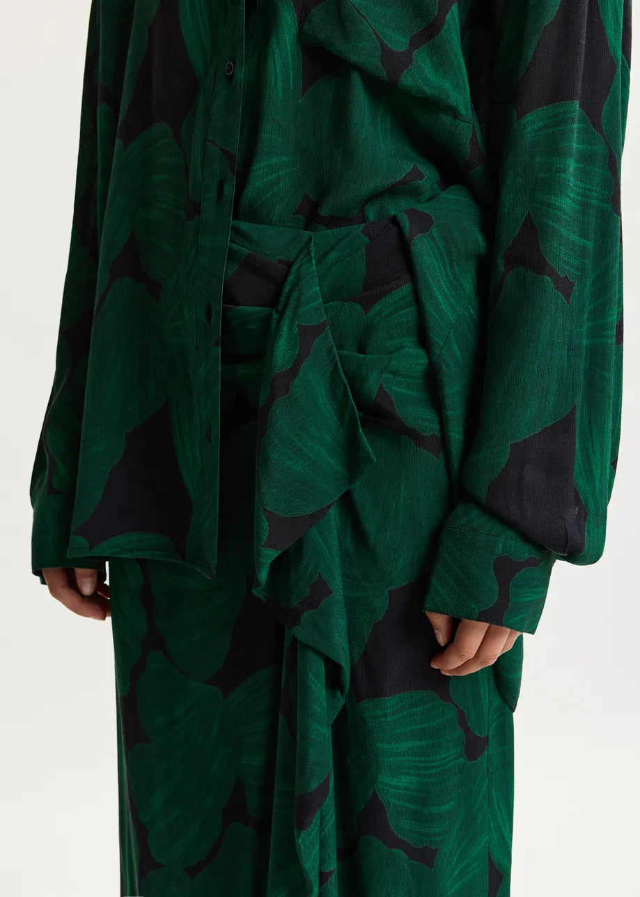 A close-up of a person wearing a long-sleeve belted shirt and matching skirt in a dark green and black abstract pattern, shown against a plain light background.