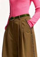 A close-up of a person wearing brown wide-leg pleated trousers with decorative black studs down the side, a green belt, and a bright pink ribbed sweater against a plain white background.