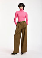A model wearing a bright pink turtleneck sweater, wide-leg brown trousers with decorative side chain detailing, a green belt, and black pointed shoes, photographed against a plain white studio background.