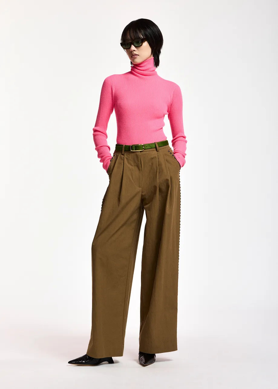 A model wearing a bright pink turtleneck sweater, wide-leg brown trousers with decorative side chain detailing, a green belt, and black pointed shoes, photographed against a plain white studio background.