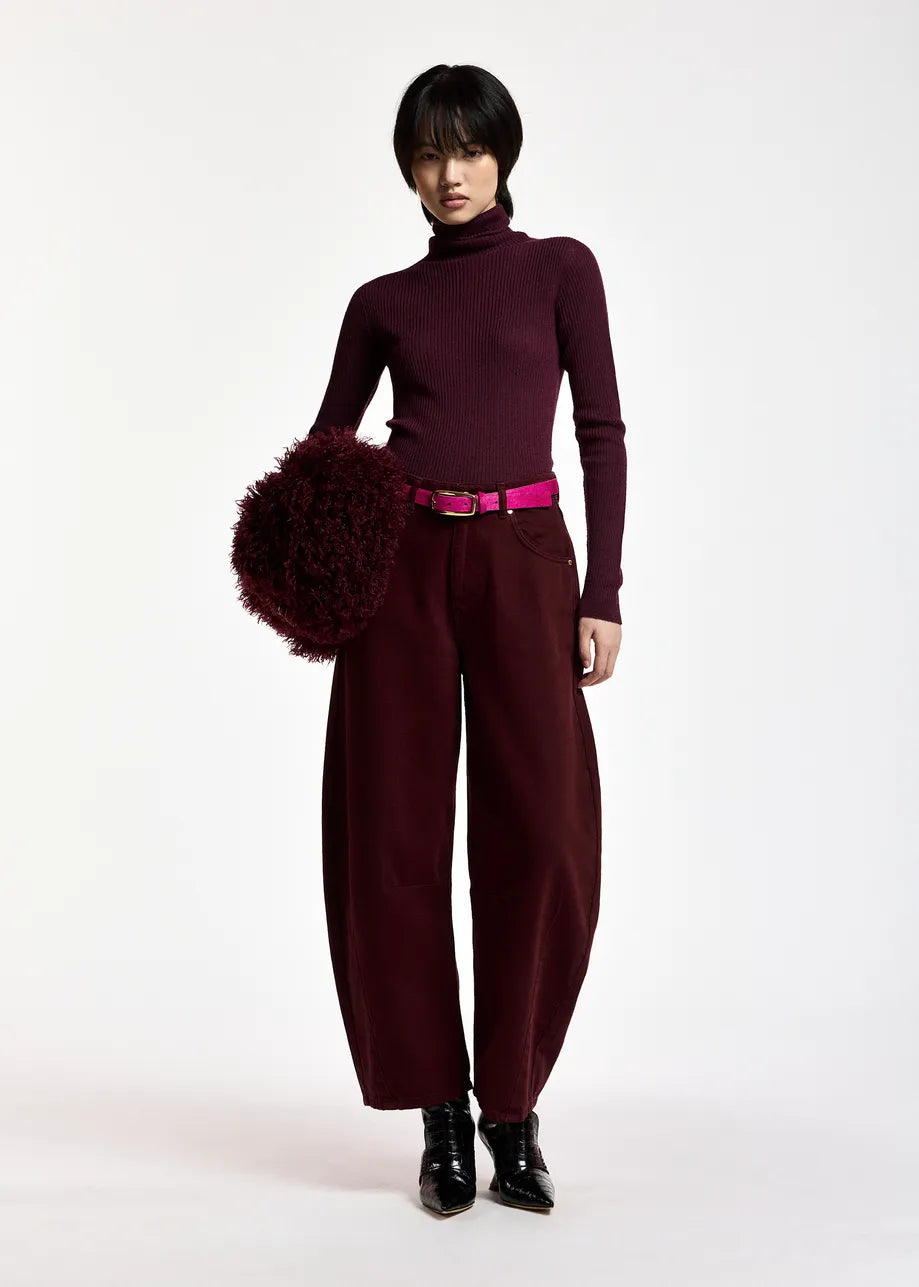 A model stands against a plain white studio background wearing a dark burgundy turtleneck sweater, matching wide-leg trousers, a bright pink belt, black pointed boots, and holding a large dark burgundy fuzzy pom-pom accessory.