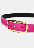 A vibrant magenta pink belt with a gold buckle, displayed horizontally on a light gray background. The inner lining is black with gold branding text.