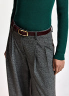 A close-up of a person wearing gray and black plaid wide-leg trousers with pleats, a dark brown leather belt with a gold buckle, and a dark green ribbed knit sweater.