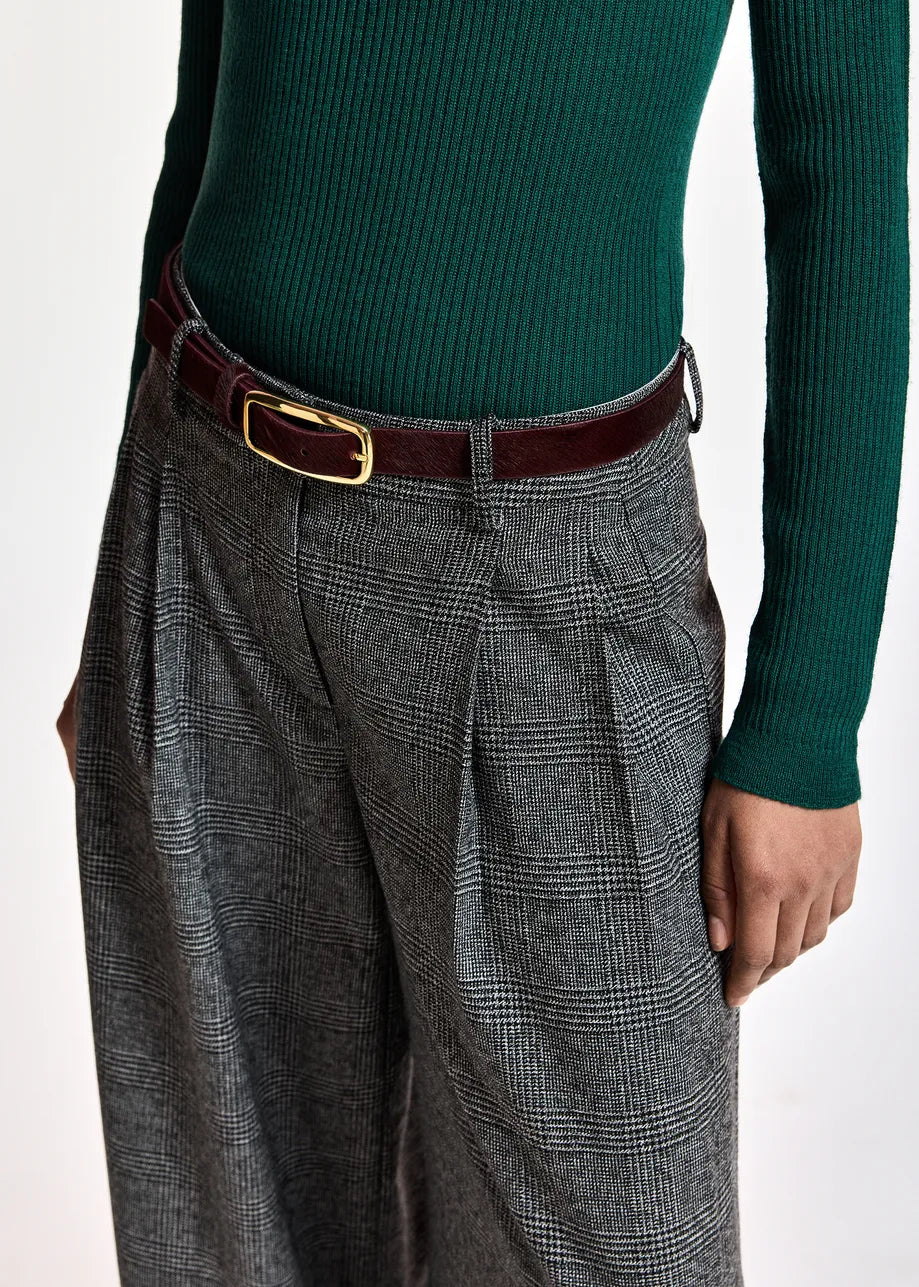 A close-up of a person wearing gray and black plaid wide-leg trousers with pleats, a dark brown leather belt with a gold buckle, and a dark green ribbed knit sweater.