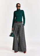 A close-up of a person wearing gray and black plaid wide-leg trousers with pleats, a dark brown leather belt with a gold buckle, and a dark green ribbed knit sweater.
