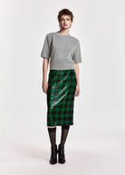 A model stands against a light gray studio background wearing a gray short-sleeve knit top, a green and black plaid sequined midi skirt, black tights, and black ankle boots.