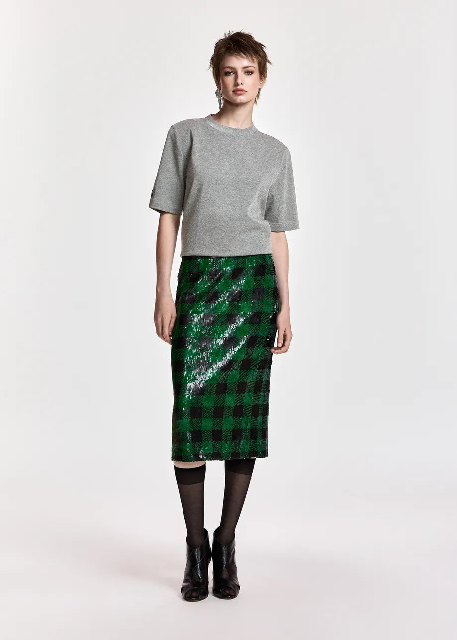 A model stands against a light gray studio background wearing a gray short-sleeve knit top, a green and black plaid sequined midi skirt, black tights, and black ankle boots.