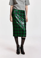 A model wearing a green and black sequined plaid midi skirt, styled with a gray t-shirt, black tights, and black ankle boots against a plain light background.