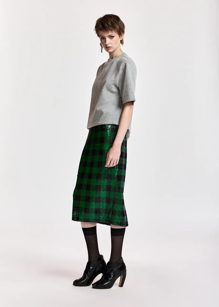 A model stands in profile wearing a light gray short-sleeve knit top, a green and black plaid midi skirt, black tights, and black high heels against a plain light gray studio background.