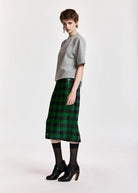 A model stands in profile wearing a light gray short-sleeve knit top, a green and black plaid midi skirt, black tights, and black high heels against a plain light gray studio background.