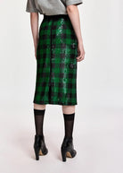 A model wearing a green and black sequined plaid midi skirt, styled with a gray t-shirt, black tights, and black ankle boots against a plain light background.