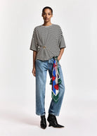 A close-up of a person wearing blue jeans and a black-and-white striped top, with a colorful patterned scarf tied around the waist over the jeans.
