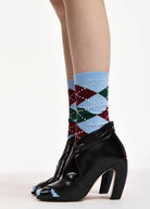 A close-up of a person's legs wearing patterned crew socks with black peep-toe high-heeled ankle boots against a plain white studio background.