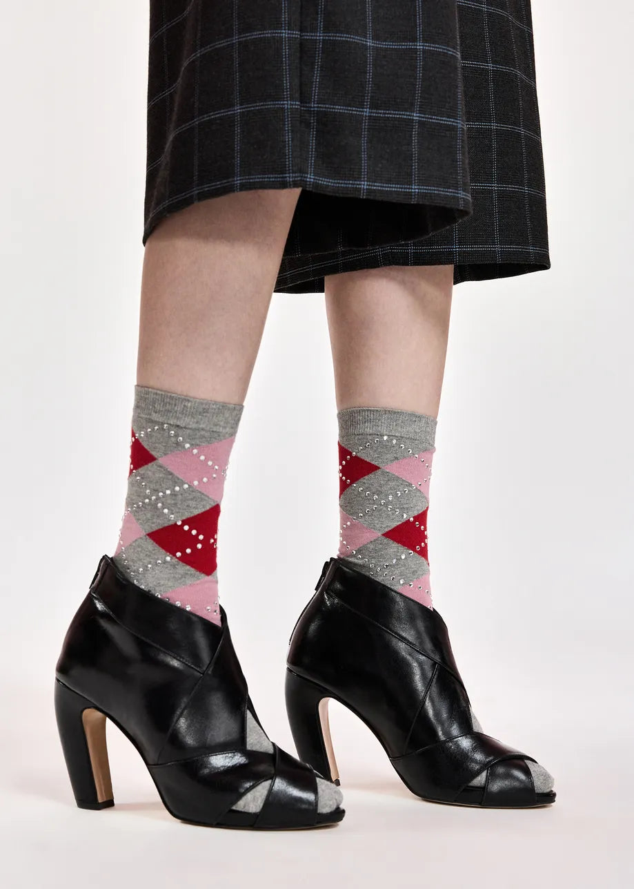 A person wearing gray argyle socks with red and pink diamonds, paired with black peep-toe high heels and a dark plaid skirt, shown from the knees down on a light studio background.