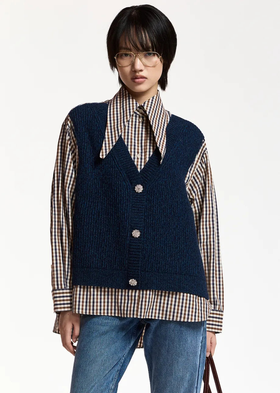 A navy blue knit vest layered over a beige, brown, and white gingham shirt, featuring a pointed collar and three silver buttons on the front, set against a plain light gray background.