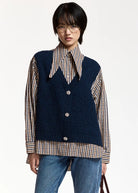 A navy blue knit vest layered over a beige, brown, and white gingham shirt, featuring a pointed collar and three silver buttons on the front, set against a plain light gray background.