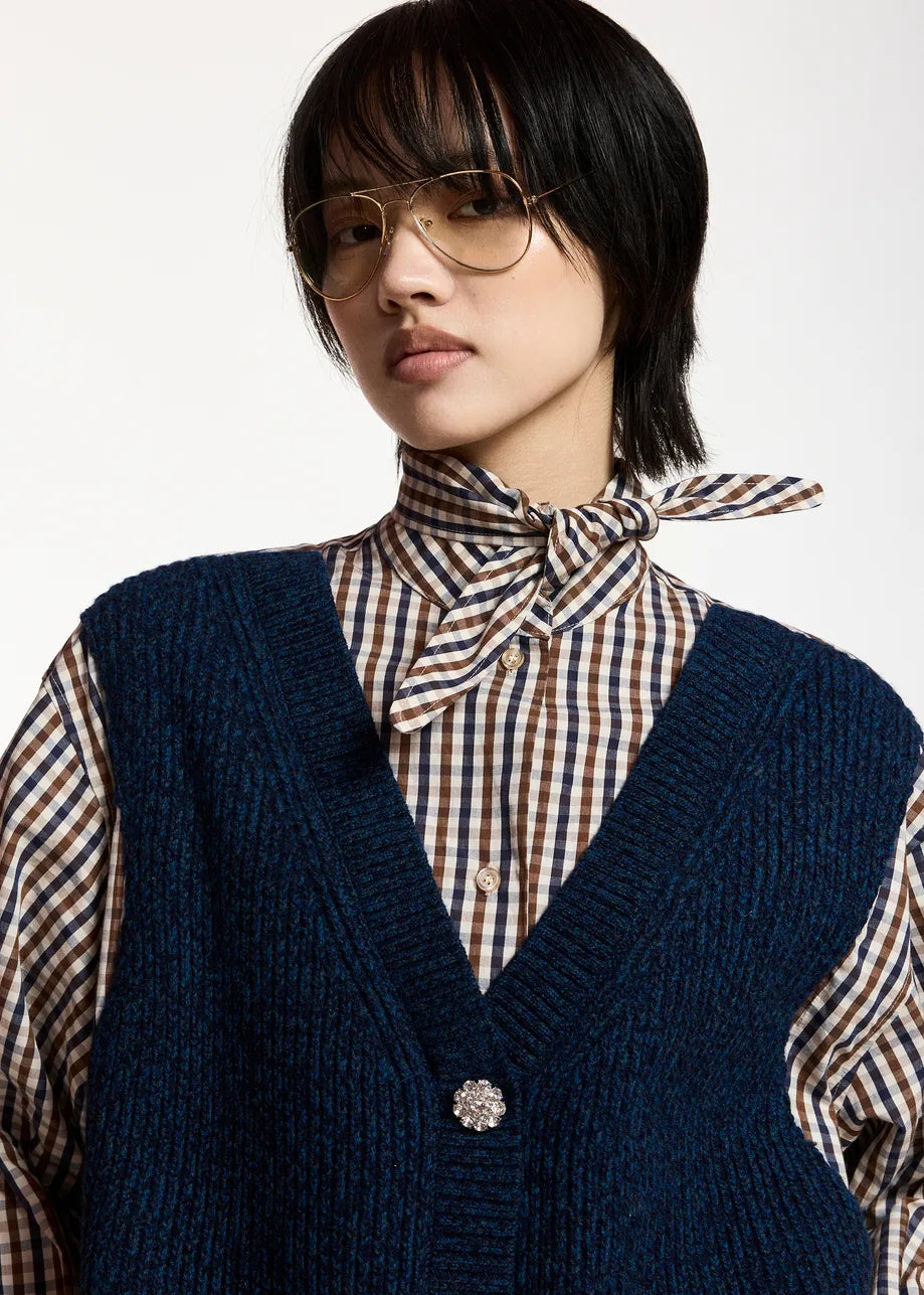 A model with short black hair and glasses wearing a navy blue ribbed knit cardigan over a brown, beige, and white gingham shirt with a matching necktie, against a plain white background.