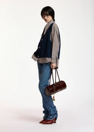 A model stands in profile wearing a navy blue vest over a beige and navy gingham shirt, blue jeans, and red pointed-toe heels, while holding a dark brown cylindrical handbag against a plain white studio background.