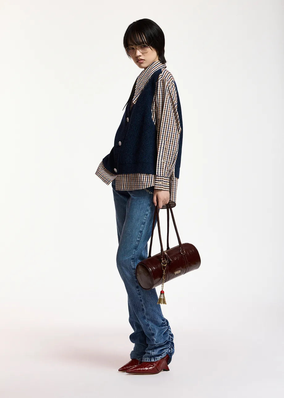 A model stands in profile wearing a navy blue vest over a beige and navy gingham shirt, blue jeans, and red pointed-toe heels, while holding a dark brown cylindrical handbag against a plain white studio background.